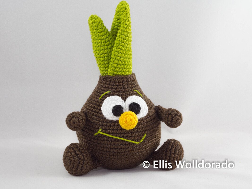 Braune gehäkelte Amigurumi-Pflanze mit zwei grünen Trieben, großen weißen Augen und gelber Nase auf hellem Hintergrund