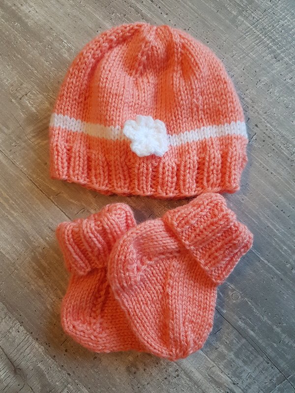 Korallfarbene gestrickte Babymütze mit weißer Blüte und passenden Söckchen auf grauem Holzuntergrund