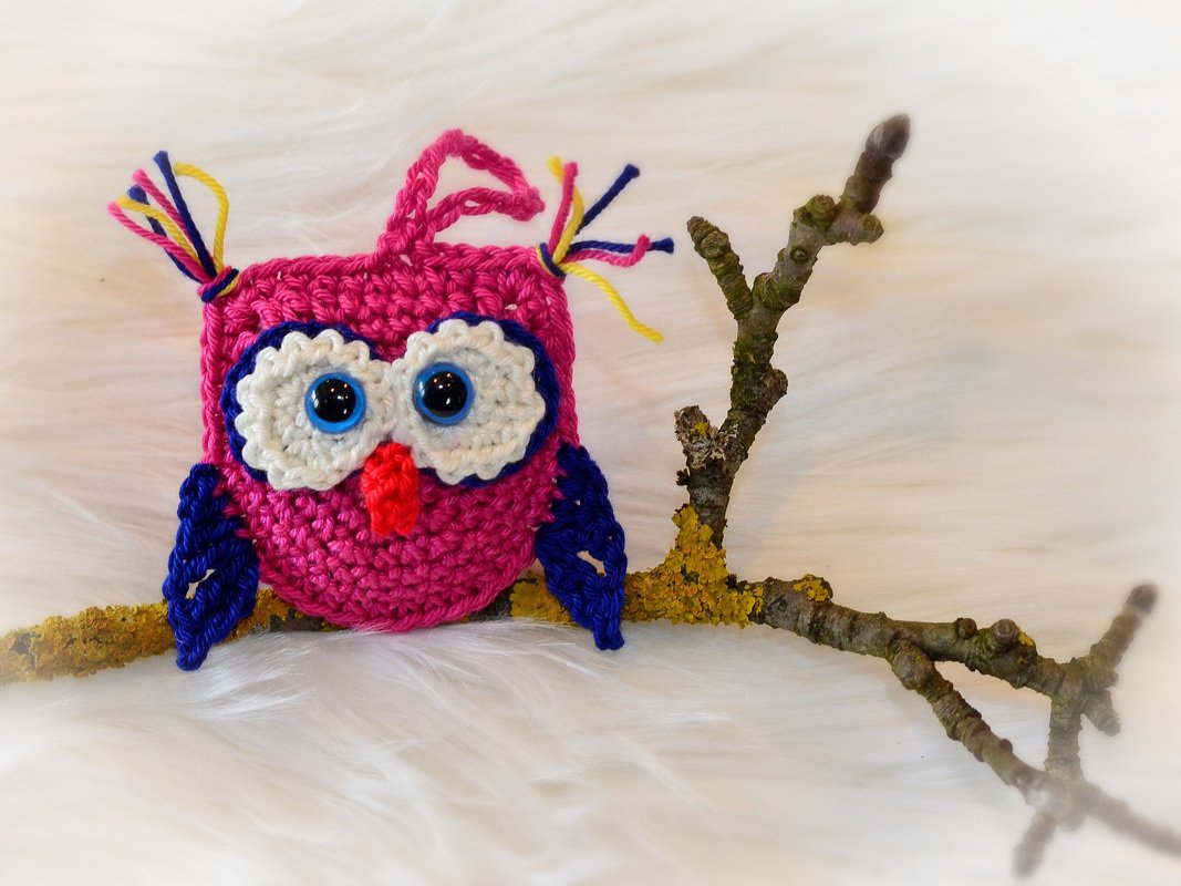 Quadratische Amigurumi-Eule in Pink mit blauen Flügeln auf einem Ast