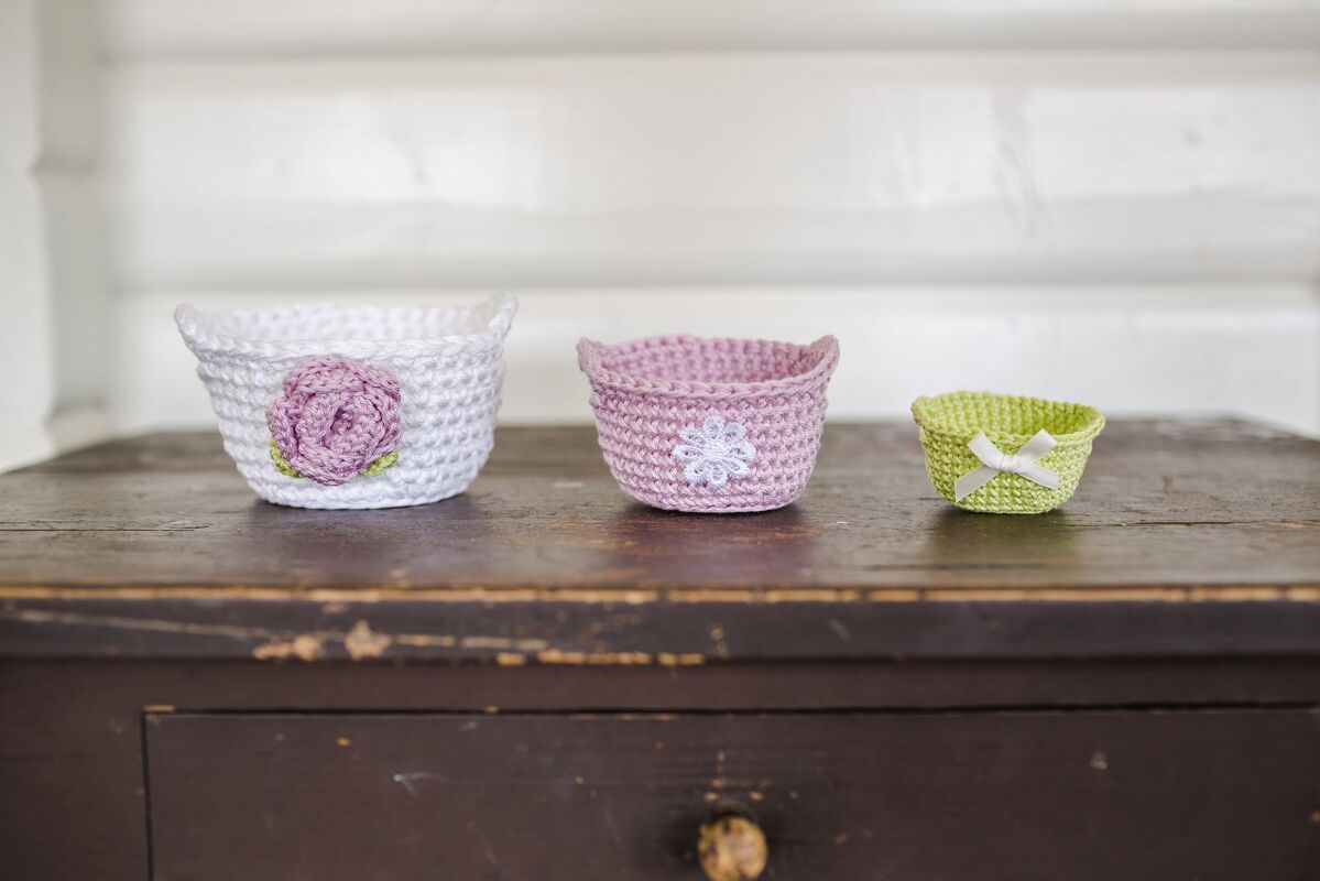 Drei kleine gehäkelte Körbchen in Weiß, Rosa und Grün auf einer dunklen Holzoberfläche