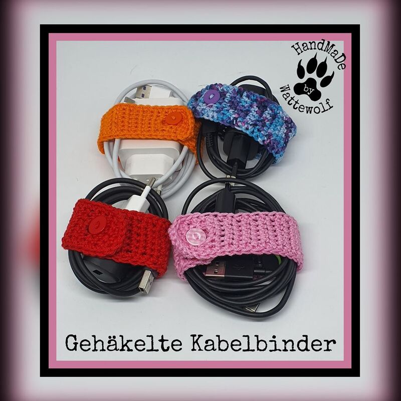 Vier gehäkelte Kabelbinder in Orange, Blau, Rot und Rosa, jeweils mit Knopf, halten aufgewickelte Ladekabel.