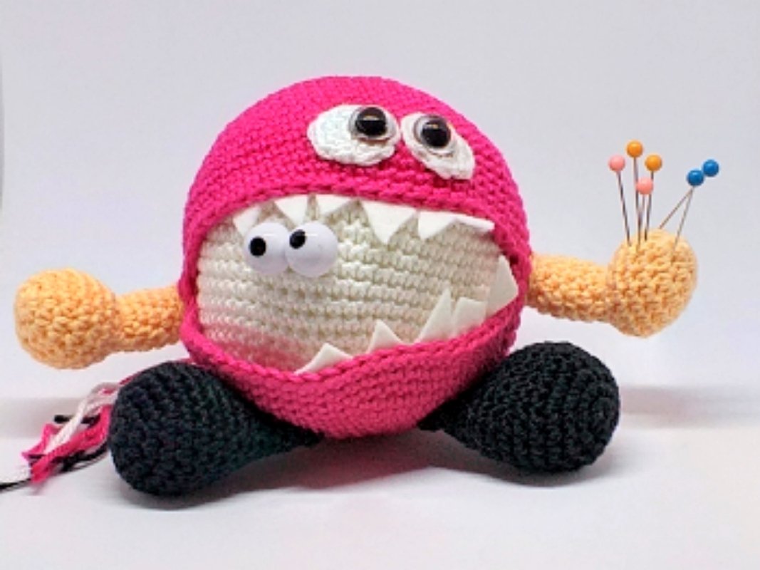 Crocheted pink round monster pincushion with white mouth, triangular teeth and colorful pins in one hand