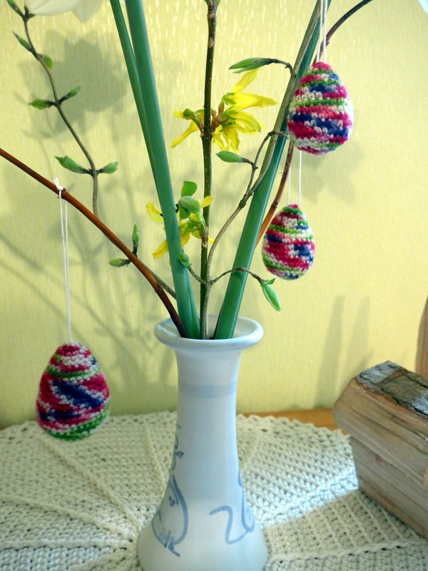 Weiße Vase mit Zweigen und drei mehrfarbigen gehäkelten Ostereiern