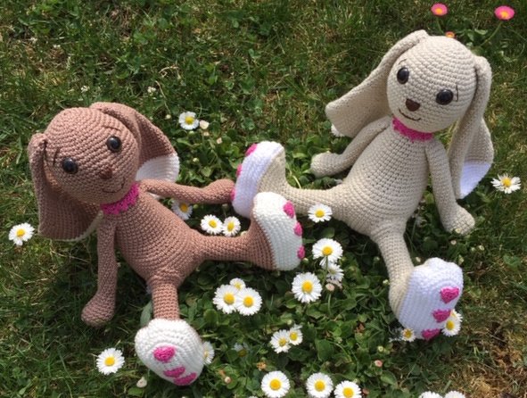Zwei gehäkelte Amigurumi-Hasen (beige und creme) mit langen Ohren und herzförmigen Fußapplikationen auf einer Wiese mit Gänseblümchen