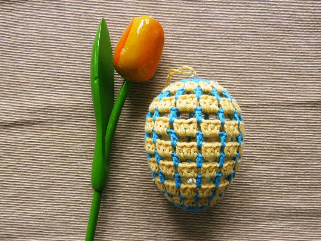 Gehäkeltes gelb-hellblaues Oster-Ei mit Gittermuster neben einer orange-gelben Holz-Tulpe auf beigem Stoff.