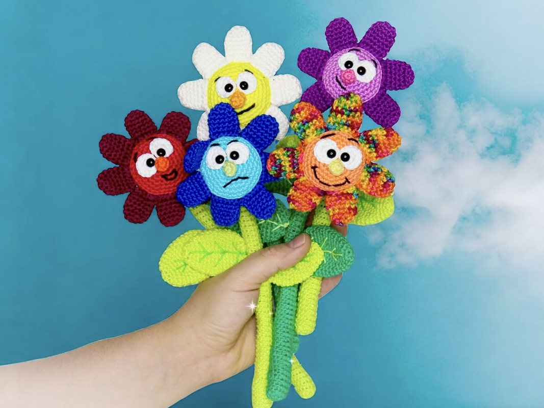 Hand holding a bouquet of five colorful crocheted amigurumi flowers with expressive faces and green crocheted stems against a blue background