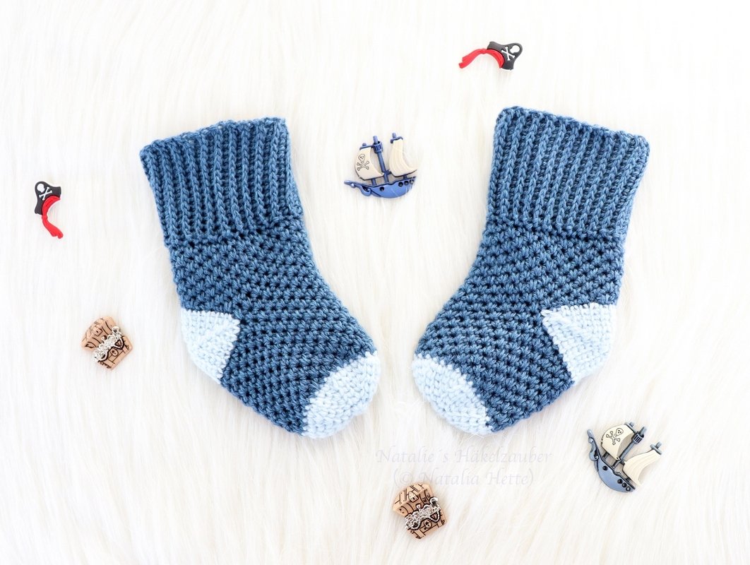 Nautical-inspired pair of blue crochet baby socks with ribbed cuffs and pale-blue toes, photographed with small ship buttons