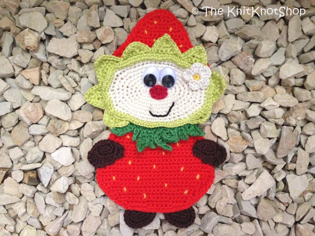 Draufsicht einer gehäkelten Erdbeer-Applikation mit Gesicht und grünen Blättern auf hellem Kies