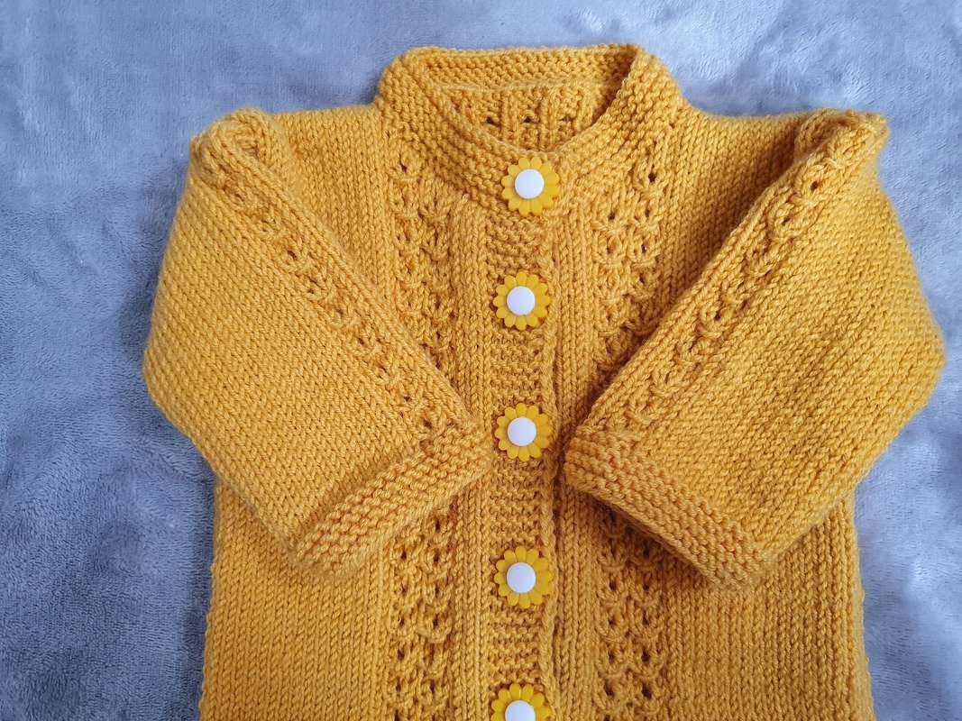 Gelbe gestrickte Babyjacke mit Blumenknöpfen und Ajourmuster auf grauem Hintergrund.