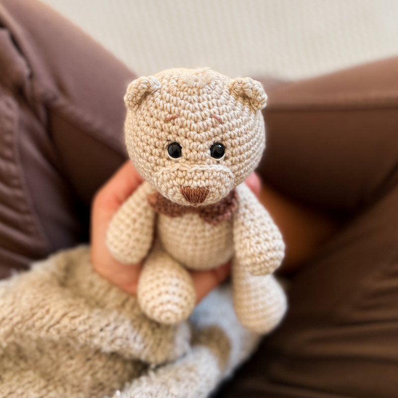 Beigefarbener handgehäkelter Teddybär mit braunem Kragen, in einer Hand gehalten