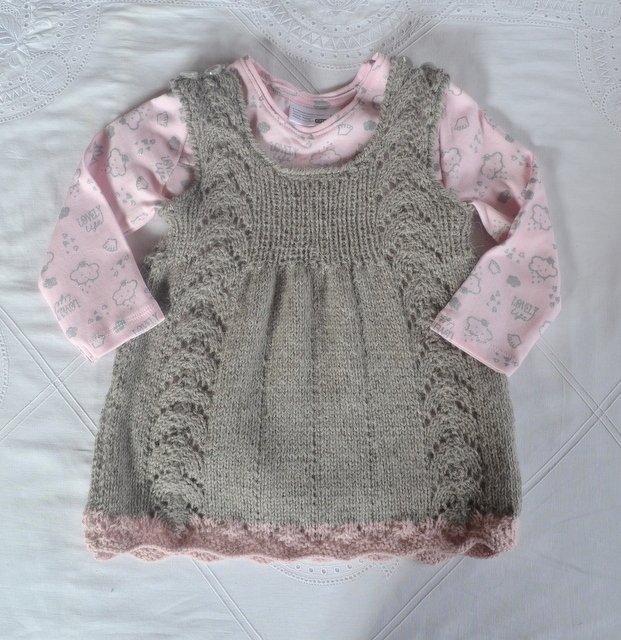 Grau gestricktes Babykleid mit Lochmuster an den Seiten und rosa gewelltem Saum, darüber ein rosa Langarmshirt mit Wolkenmotiven