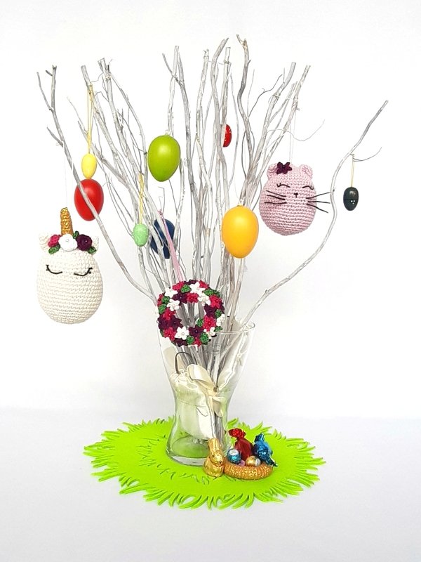 Vase mit weiß lackierten Zweigen, daran zahlreiche gehäkelte Osteranhänger (Einhorn, Katzenkopf, Blumenkranz) in Pastell- und kräftigen Farben; grüner Filzuntersetzer mit kleinen Schokoladenfiguren.