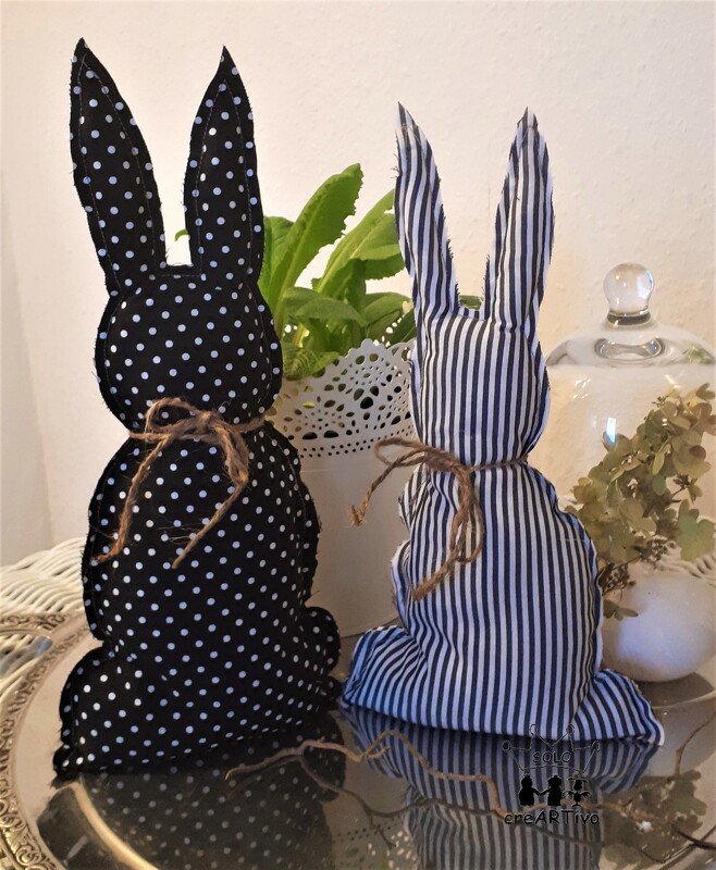 Zwei genähte Stoffhasen, einer schwarz mit weißen Punkten, einer schwarz-weiß gestreift, jeweils mit Juteschleife auf Tablett neben Pflanze