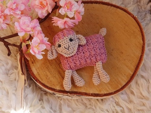 Gehäkeltes Amigurumi-Lamm mit altrosafarbenem Körper auf einer Holzscheibe neben rosa Blüten