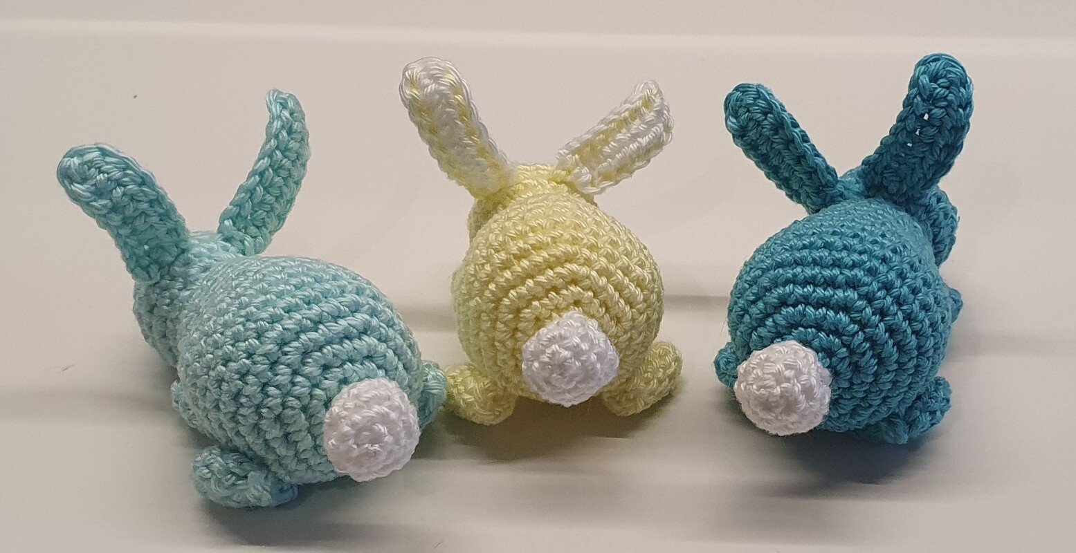 Drei gehäkelte Amigurumi‑Hasen (hellblau, gelb, türkis), Rückenansicht, weiße Pompon‑Schwänzchen