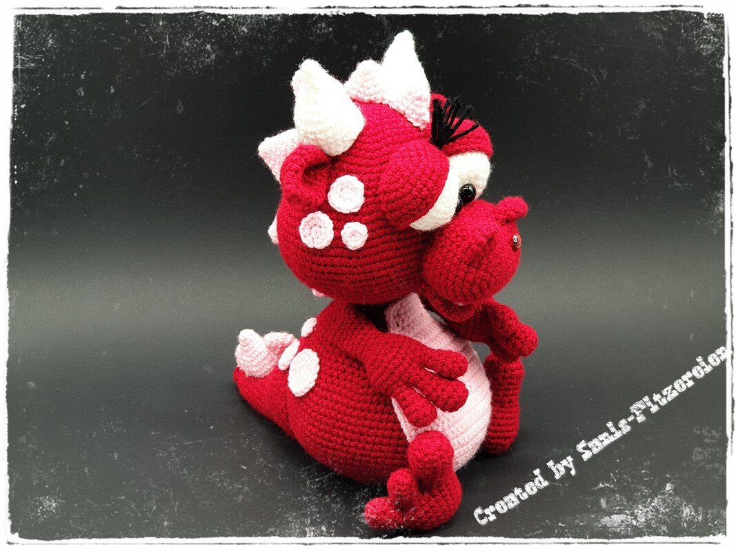 Roter gehäkelter Amigurumi-Drache sitzend mit rosa Bauch und weißen Stacheln