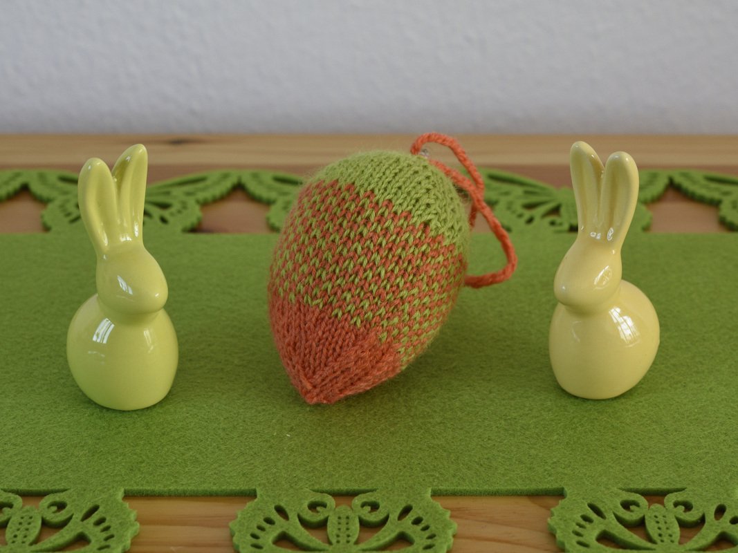 Gestricktes Ei zwischen zwei hellgelben Hasenfiguren auf grünem Filz