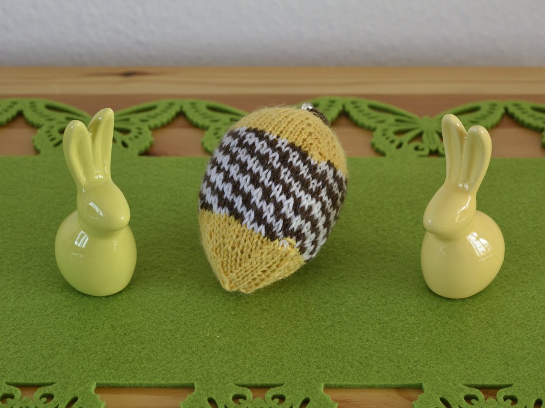 Gestricktes Osterei in Gelb, Braun und Weiß zwischen zwei gelben Keramikhasen auf grünem Filz