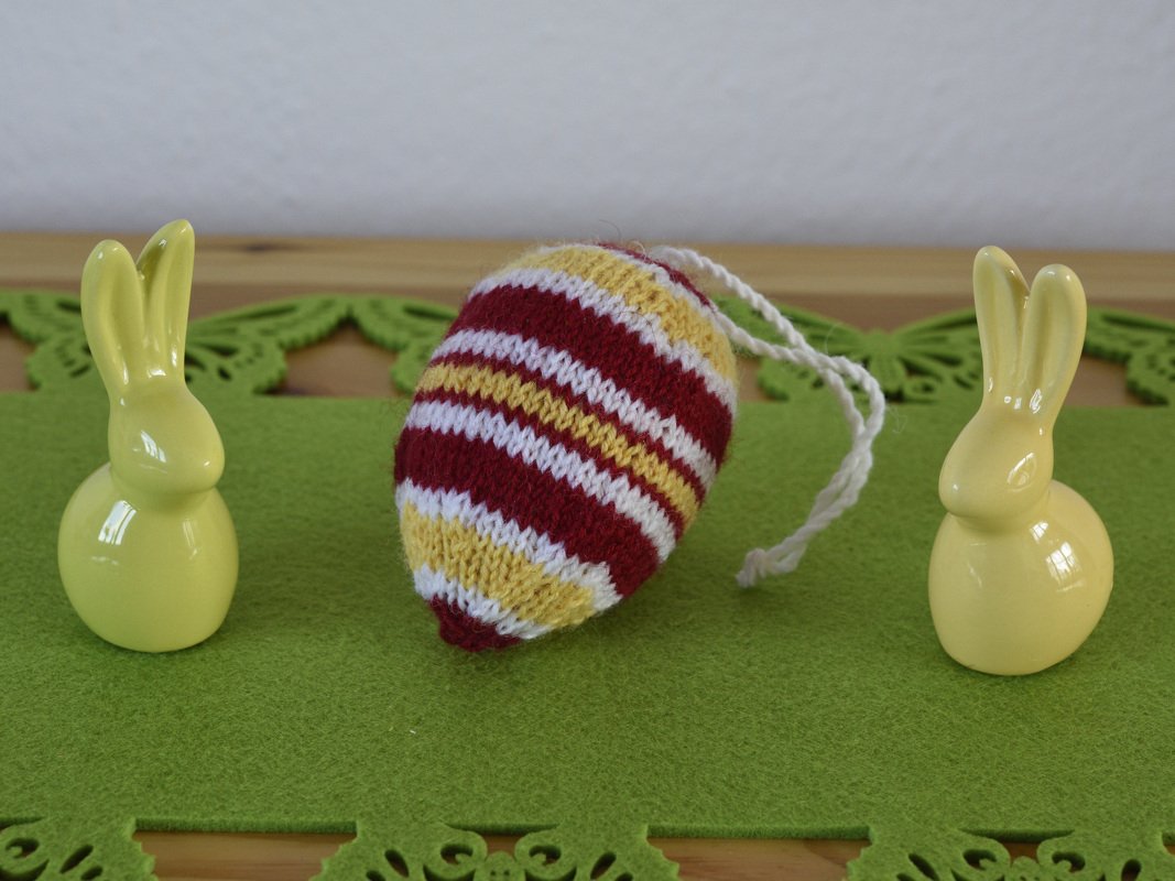 Gestricktes gestreiftes Osterei in Rot, Weiß und Gelb mit weißer Schnur, flankiert von zwei gelben Keramikhasen auf grüner Filzmatte