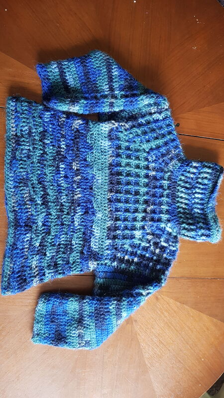 Gehäkelter Kinderpullover in Blau- und Türkistönen, liegend auf einer Holzoberfläche; Häkelstrukturen gut sichtbar.