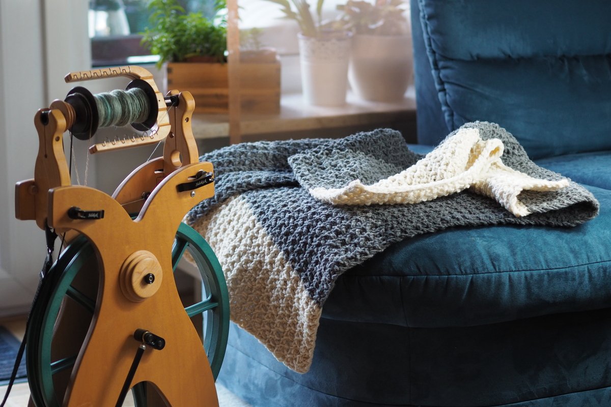 Zweifarbige gestrickte Babydecke (Grau & Creme) mit markanter Maschenstruktur auf blauem Sofa, Spinnrad mit Garn daneben