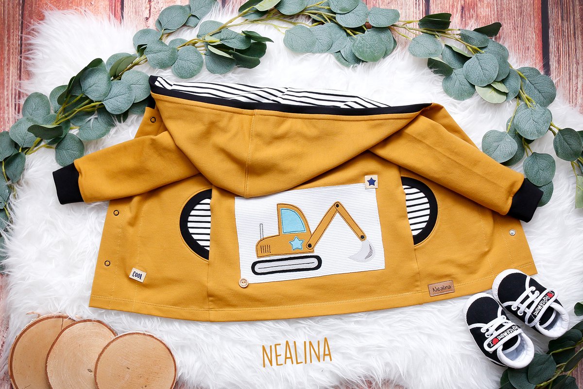 Detailaufnahme einer senfgelben Kapuzenjacke mit zentraler Bagger-Applikation, runden Taschenöffnungen und gestreiftem Futter