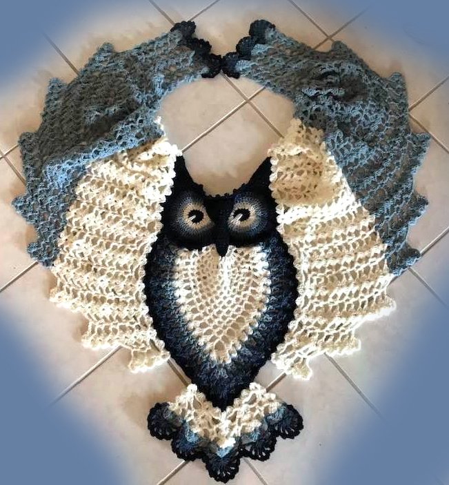 Crocheted owl-shaped triangle shawl with openwork wings in blue and cream, laid flat on tiles.