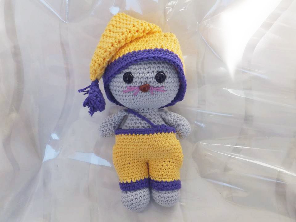 Gehäkelte graue Amigurumi-Maus mit gelber Zipfelmütze und gelber Latzhose, lila Details, auf hellem Hintergrund.