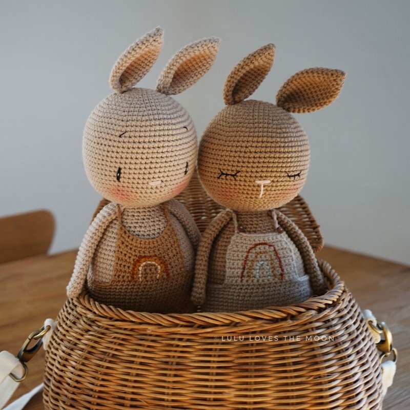 Zwei gehäkelte Amigurumi-Hasen mit Regenbogen-Overalls in einem Weidenkorb.