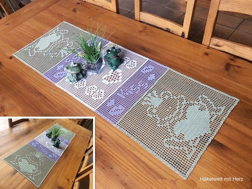 Gehäkelter Tischläufer mit Herz- und Blockmustern in Pastellgrün, Weiß und Lila auf einem Holztisch, dekoriert mit Froschfiguren und einer kleinen Pflanze.