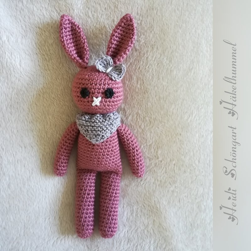 Rosa gehäkelter Amigurumi-Hase mit grauer Schleife am Ohr und grauem Schal auf hellem, flauschigem Untergrund.