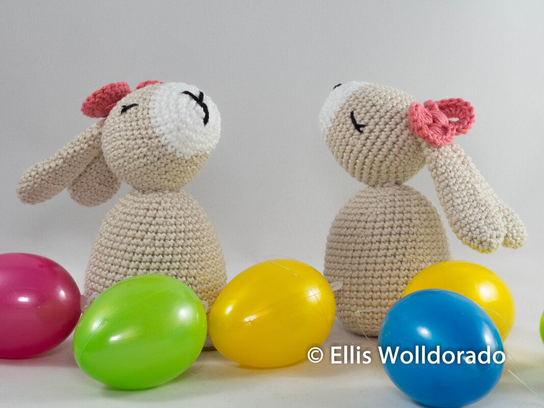 Zwei Amigurumi-Hasen in hellem Beige mit rosa Schleifen, sitzend neben bunten Ostereiern.