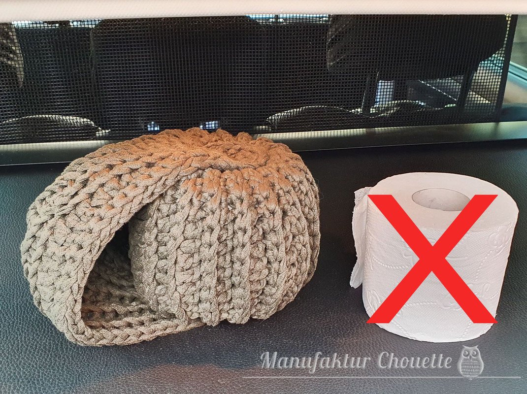 Beigefarbene, grob gehäkelte Hülle neben einer Toilettenpapierrolle mit rotem X