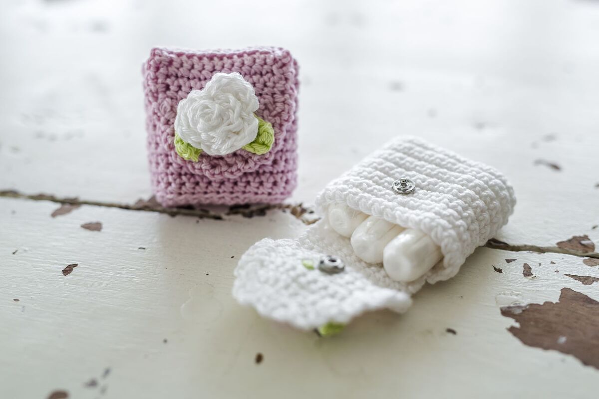 Zwei gehäkelte Täschchen: ein rosa mit weißer Häkelrose, das andere weiß und geöffnet mit drei Tampons, auf einem alten Holztisch.