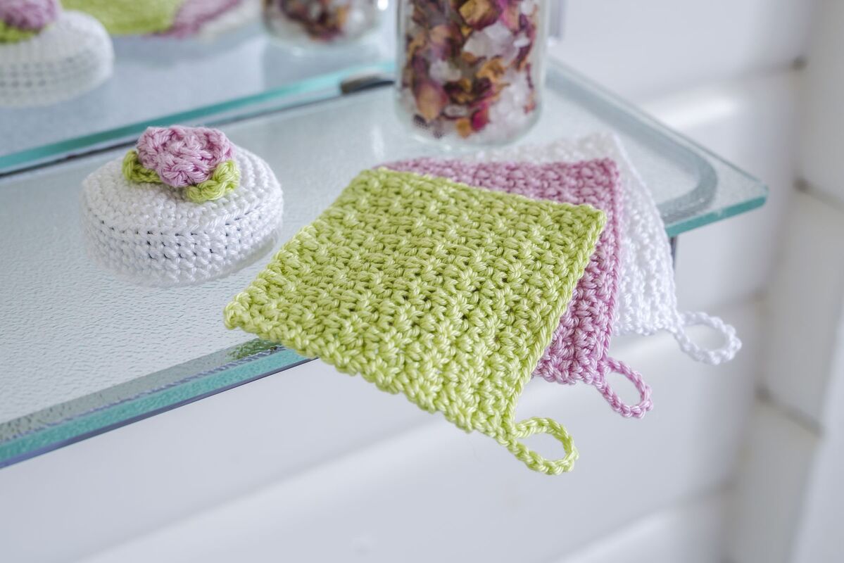 Drei schnell gehäkelte quadratische Mini-Pads in Pastellgrün, Rosa und Weiß mit kleinen Schlaufen auf einer Glasablage