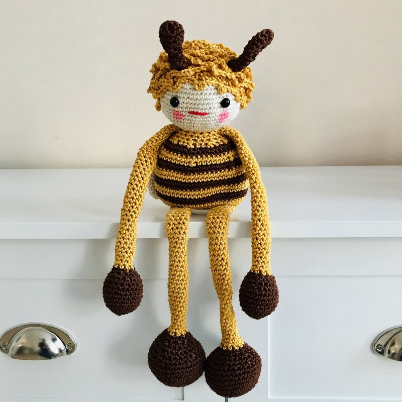 Gelb-braun gestreiftes gehäkeltes Amigurumi mit lockigem Haar, Antennen und langen Armen und Beinen, sitzend auf einer weißen Kommode
