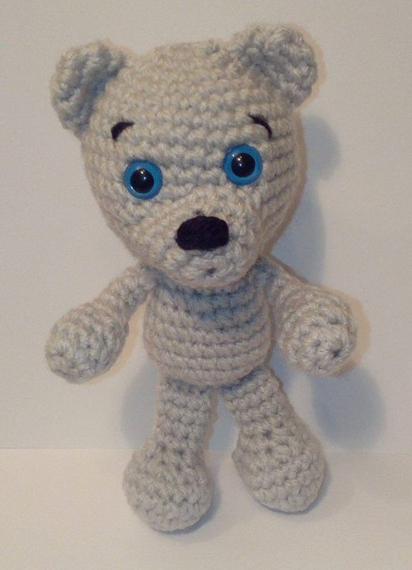 Handgehäkelter hellgrauer Amigurumi-Teddybär mit blauen Augen und schwarzer Nase, stehend.
