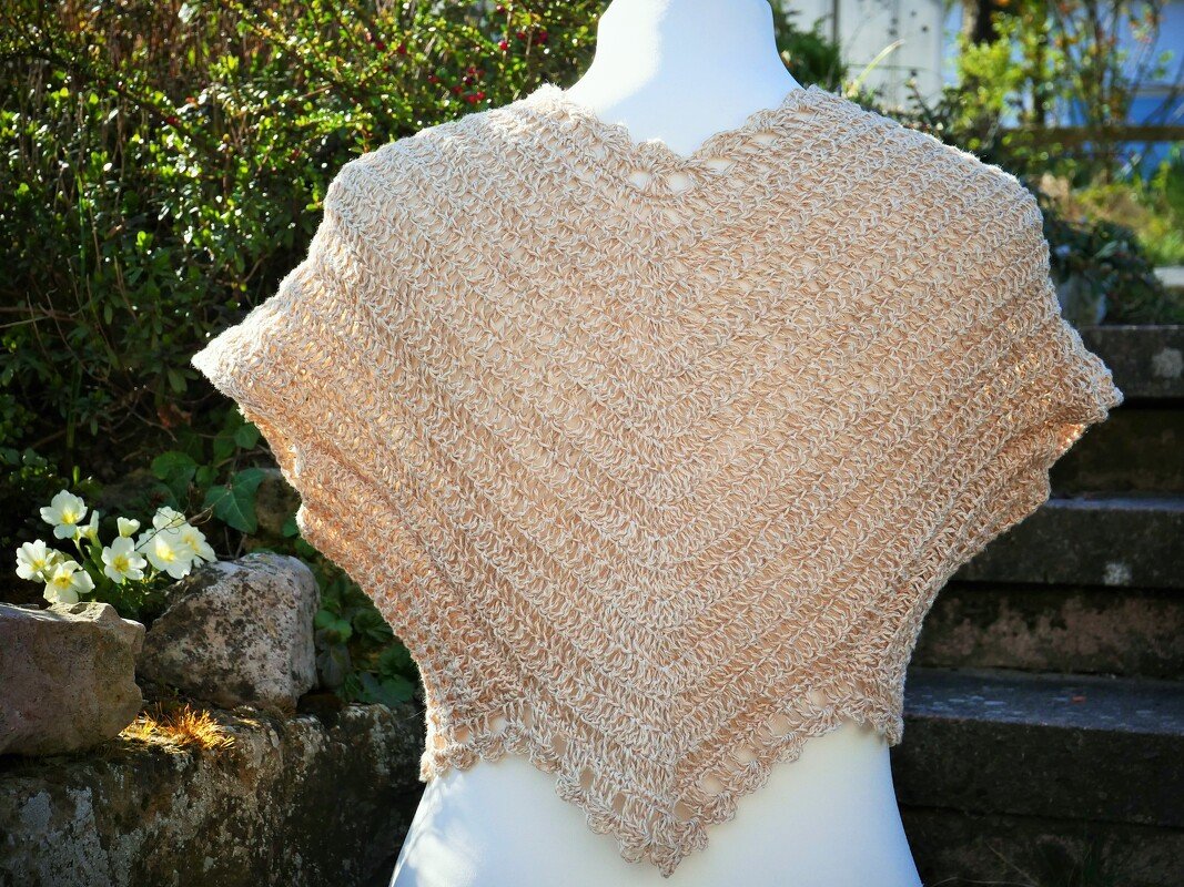 Beiges gehäkeltes Schultertuch mit Lochmuster auf weißer Schaufensterpuppe vor Pflanzenkulisse.