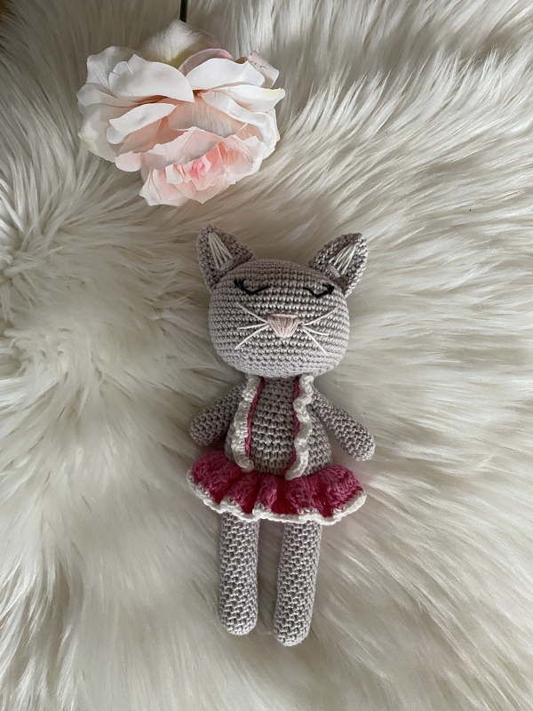 Gehäkelte graue Amigurumi‑Katze mit rosa Tüllrock und weißem Oberteil auf weißem Fellimitat neben einer rosa Kunstblume