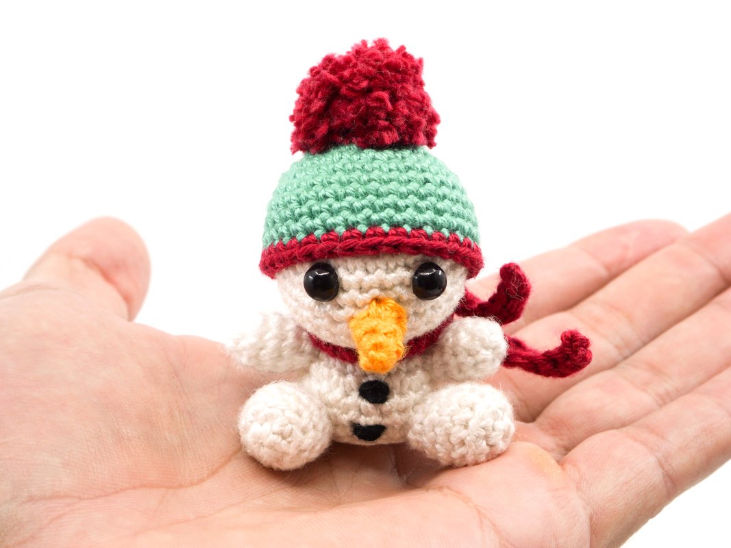Palm-sized crocheted snowman amigurumi with green hat, red pom-pom and red scarf resting in an open hand