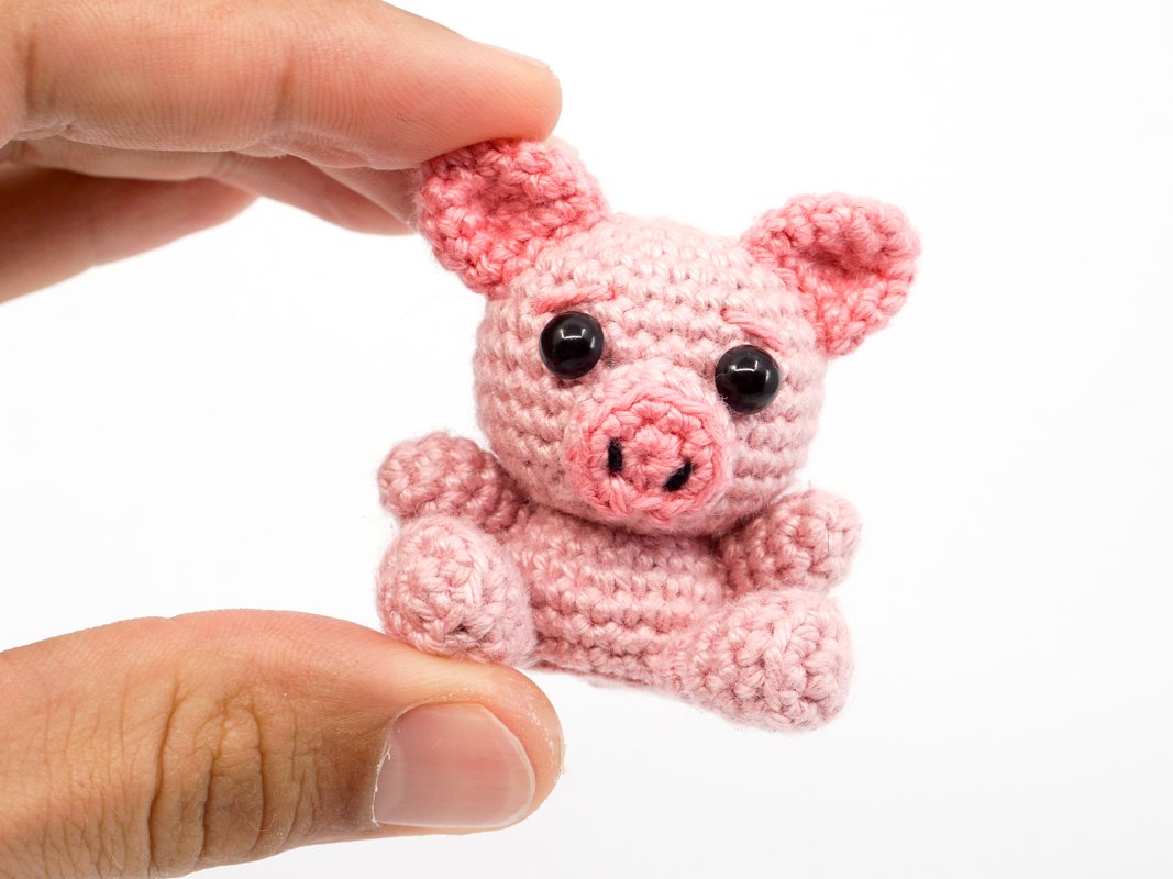 Tiny pink crocheted pig amigurumi held between thumb and index finger on a white background.