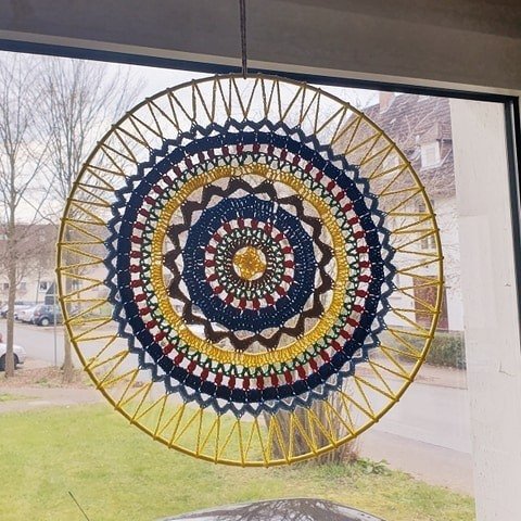 Rundes gehäkeltes Mandala in Gelb, Dunkelblau, Rot und Braun, aufgehängt im Fenster