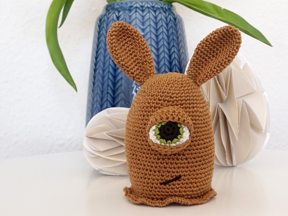 Braunes gehäkeltes Amigurumi mit Hasenohren und grünem Zyklopenauge vor blauer Vase und weißer Papierdeko.