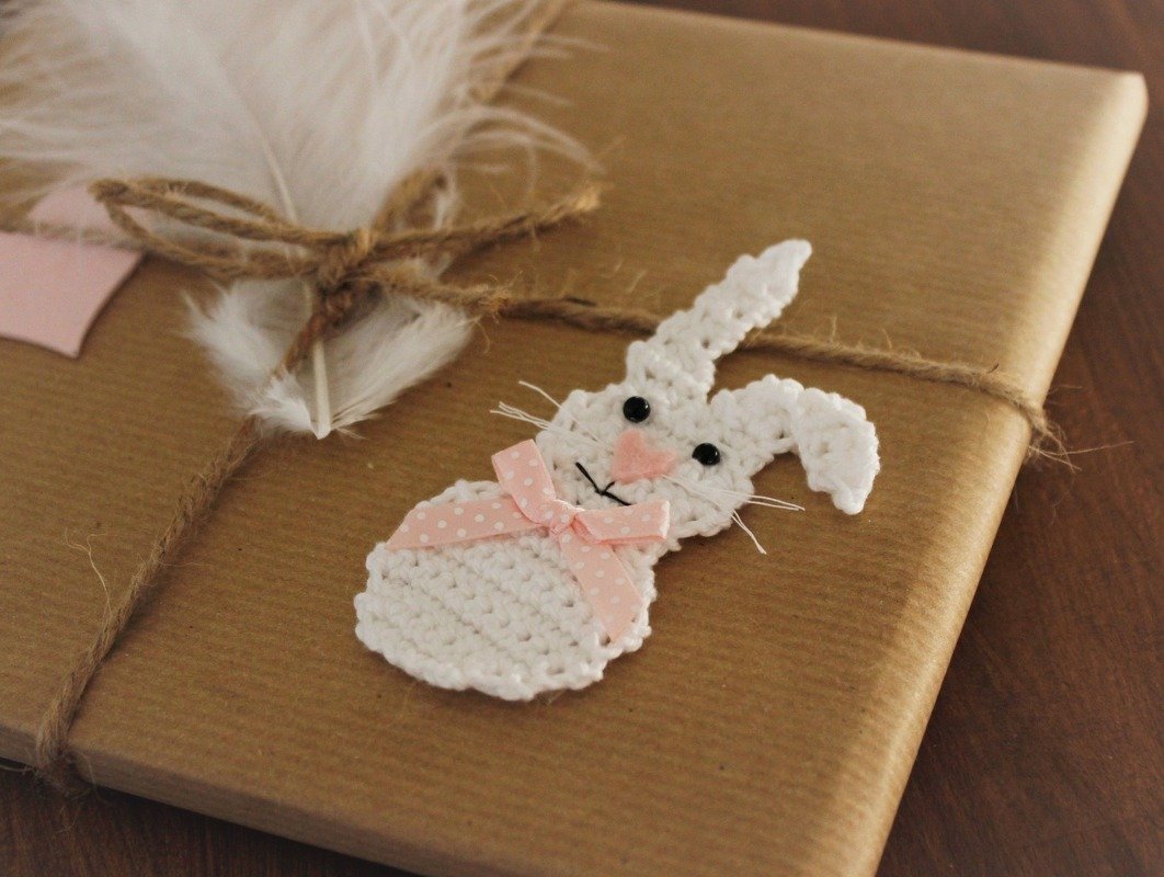 Weiße gehäkelte Hasen-Applikation mit rosa Schleife und Pompon‑Nase auf braunem Packpapier und Kordel.