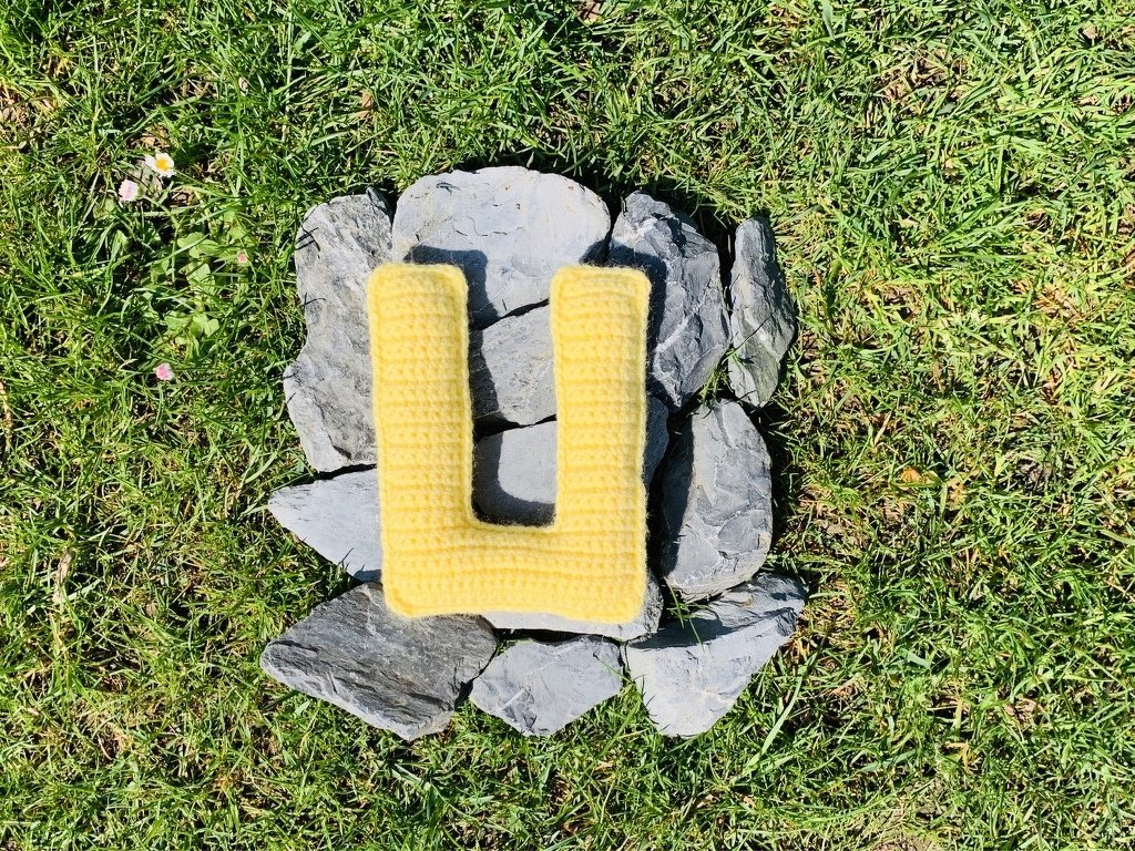 Gelber, gehäkelter Buchstabe U auf grauen Steinen im grünen Gras.