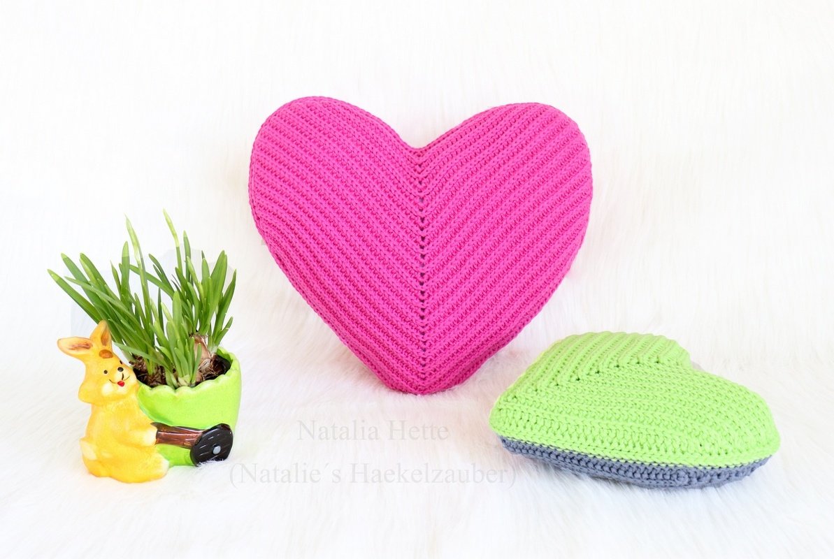 Pink crocheted heart pillow with green crocheted accent, potted plant and yellow bunny figurine on a white surface.