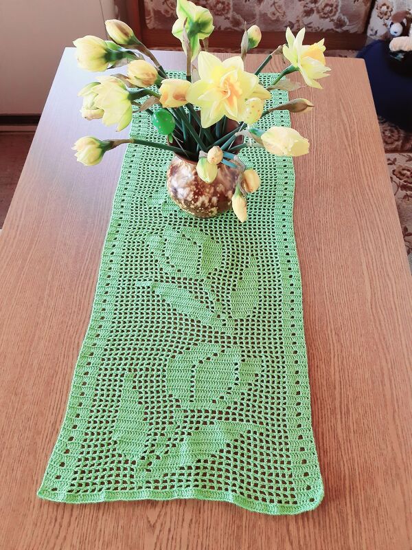 Grüner gehäkelter Tischläufer im Filetmuster mit Blumenmotiv auf Holztisch, braune Vase mit gelben Narzissen