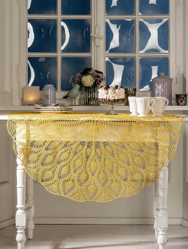 Gehäkelter gelber Tischläufer mit Fächermuster auf weißem Tisch, mit Kuchen, Blumen und Geschirr arrangiert