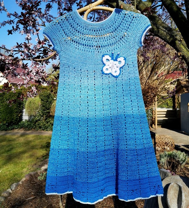 Blaues gehäkeltes Kinderkleid mit weißer Schmetterlings-Applikation an Kleiderbügel vor Garten