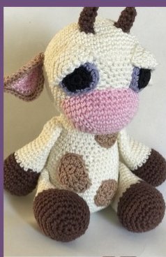 Gehäkelte sitzende Amigurumi-Kuh in Cremeweiß mit braunen Flecken und rosa Schnauze